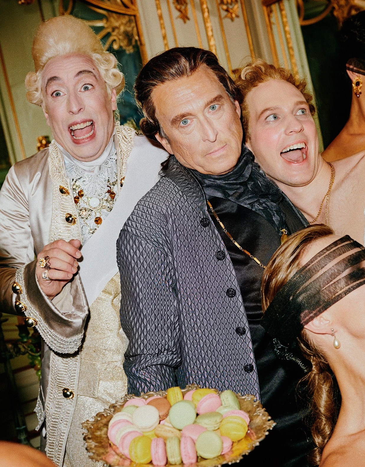 En excentrisk scen med personer klädda i historiska kläder, där några bär ögonbindlar och andra ser chockerade eller glada ut. Färgglada macarons serveras på ett fat.