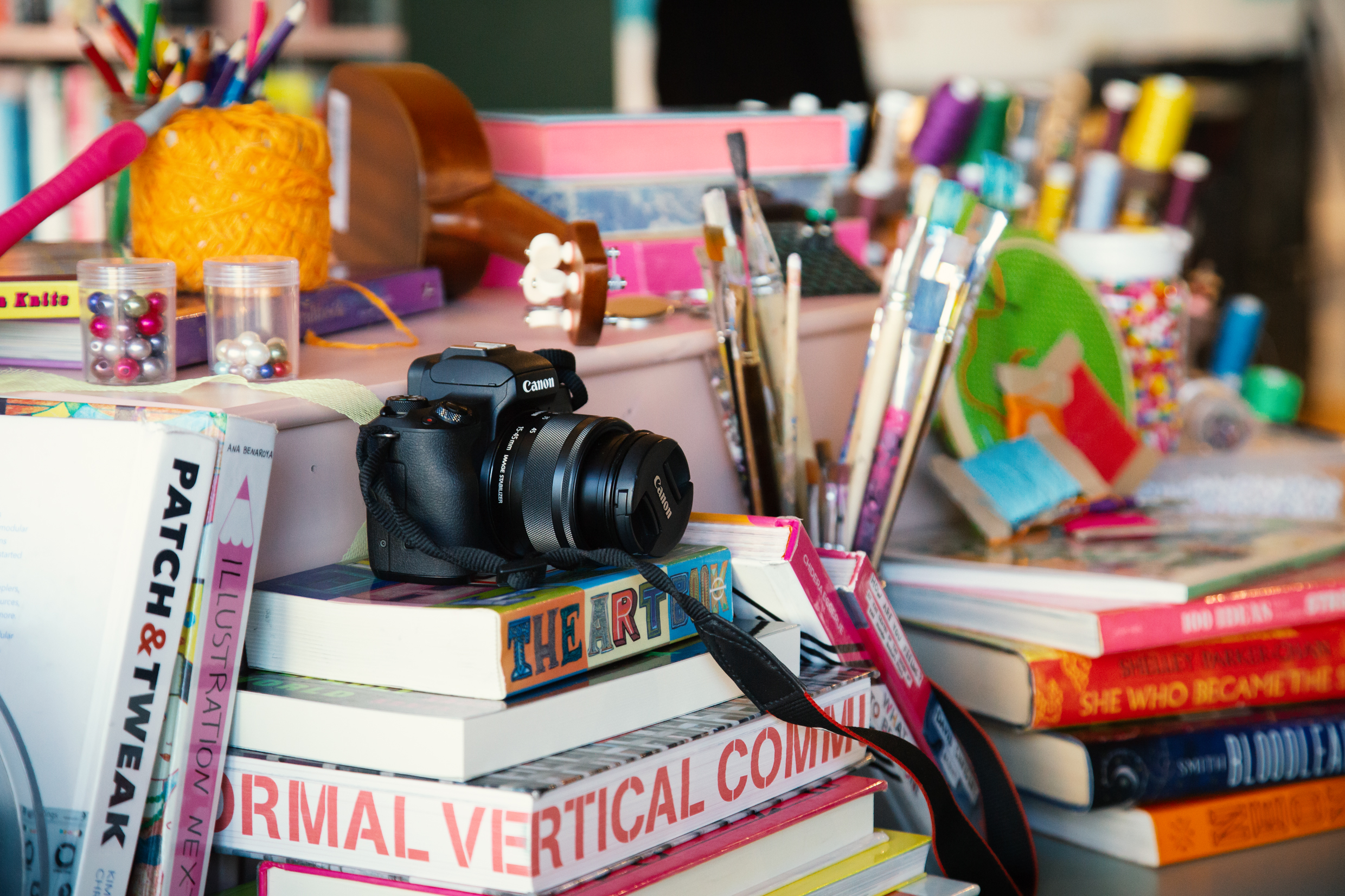 Närbild av kamera, penslar, garn och staplade böcker på ett bord.