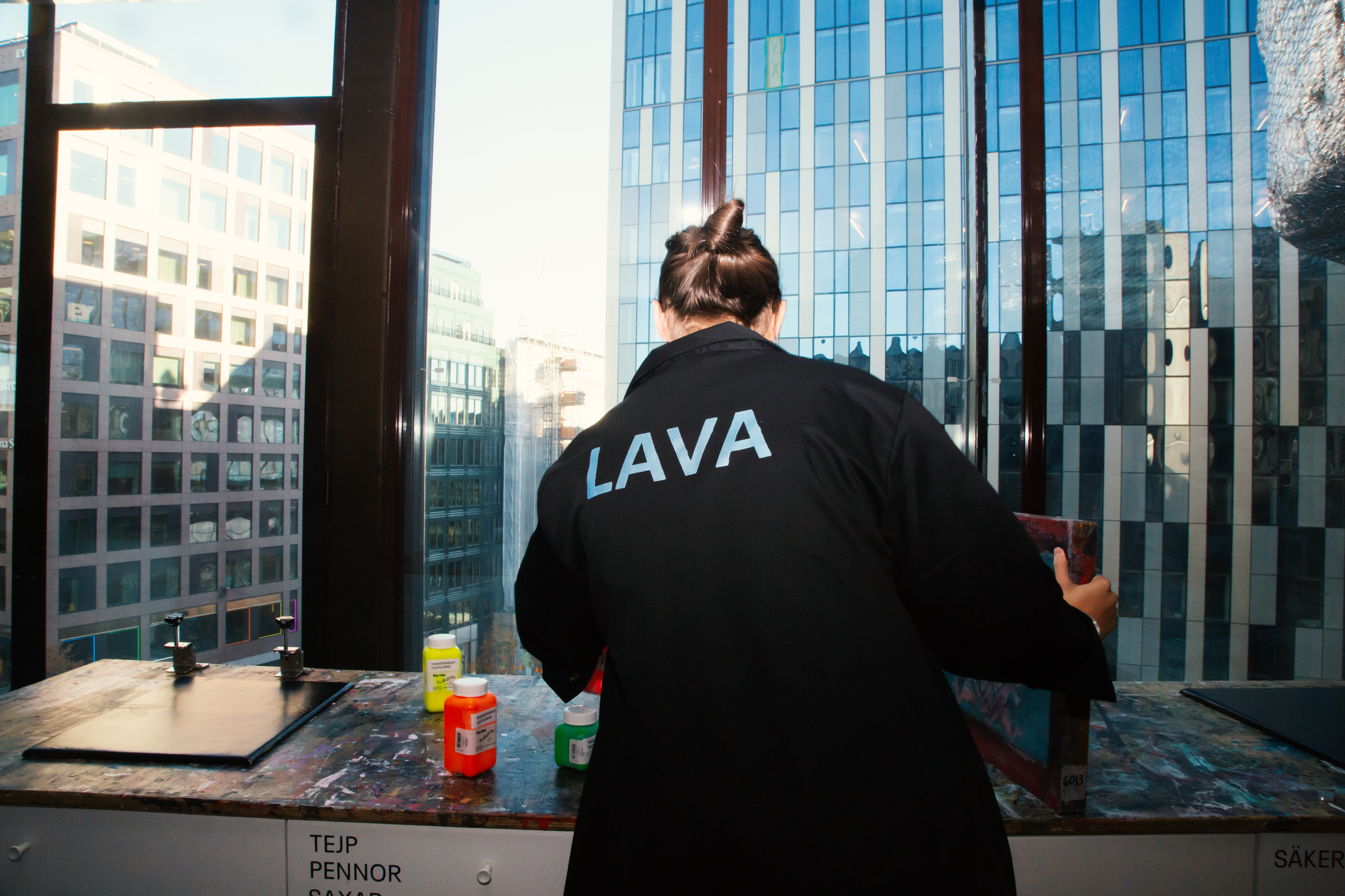 Person i svart jacka med texten "LAVA" målar vid ett bord med fönsterutsikt över stad.