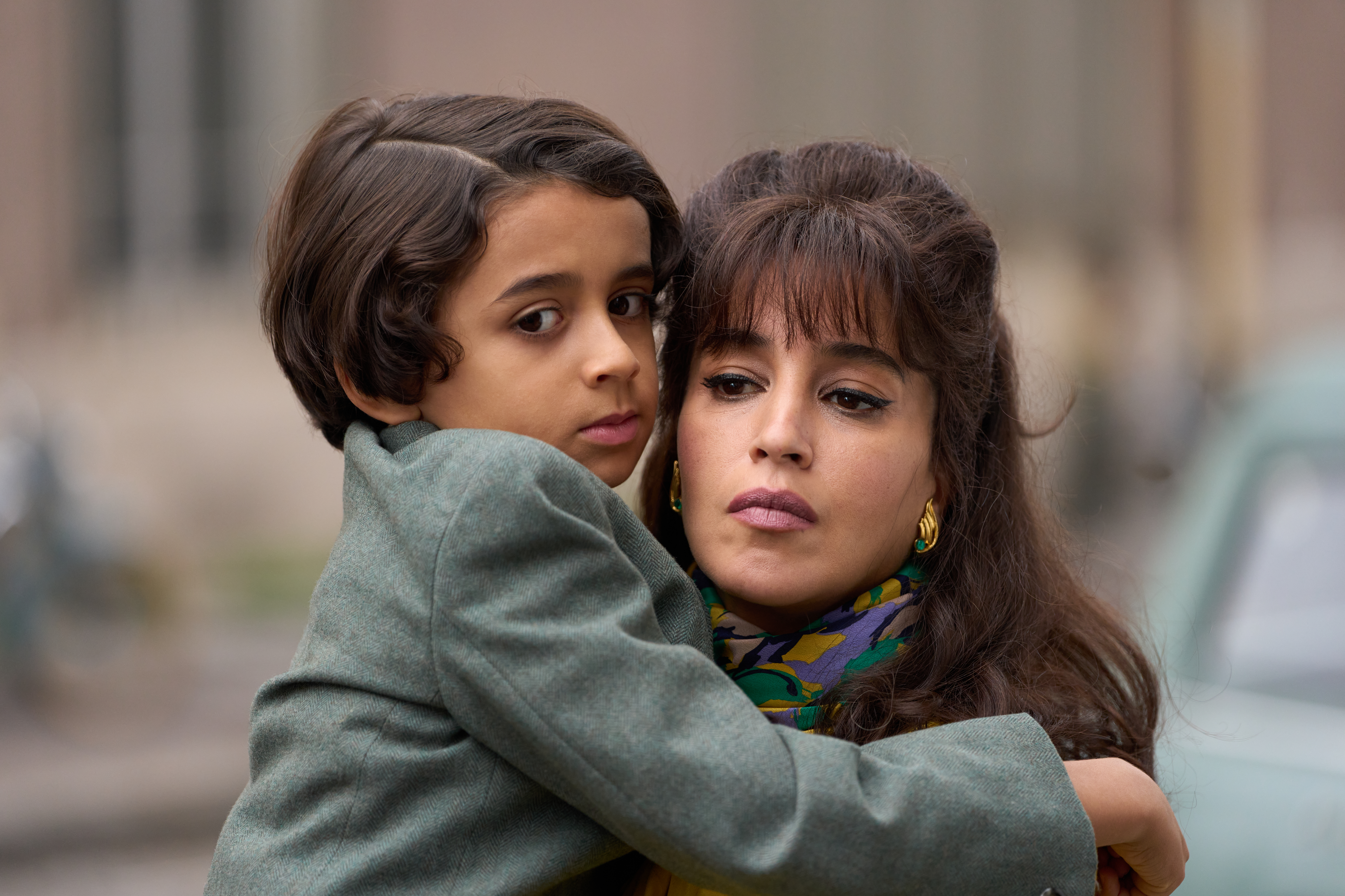 En kvinna med mörkt hår och färgglad scarf håller ett barn i famnen. Barnet, som har kort lockigt hår och bär en grå kappa, vilar huvudet mot hennes axel och tittar mot kameran. Kvinnan har en allvarlig min och bär guldörhängen. Bakgrunden är suddig och visar en gata med en bil.
