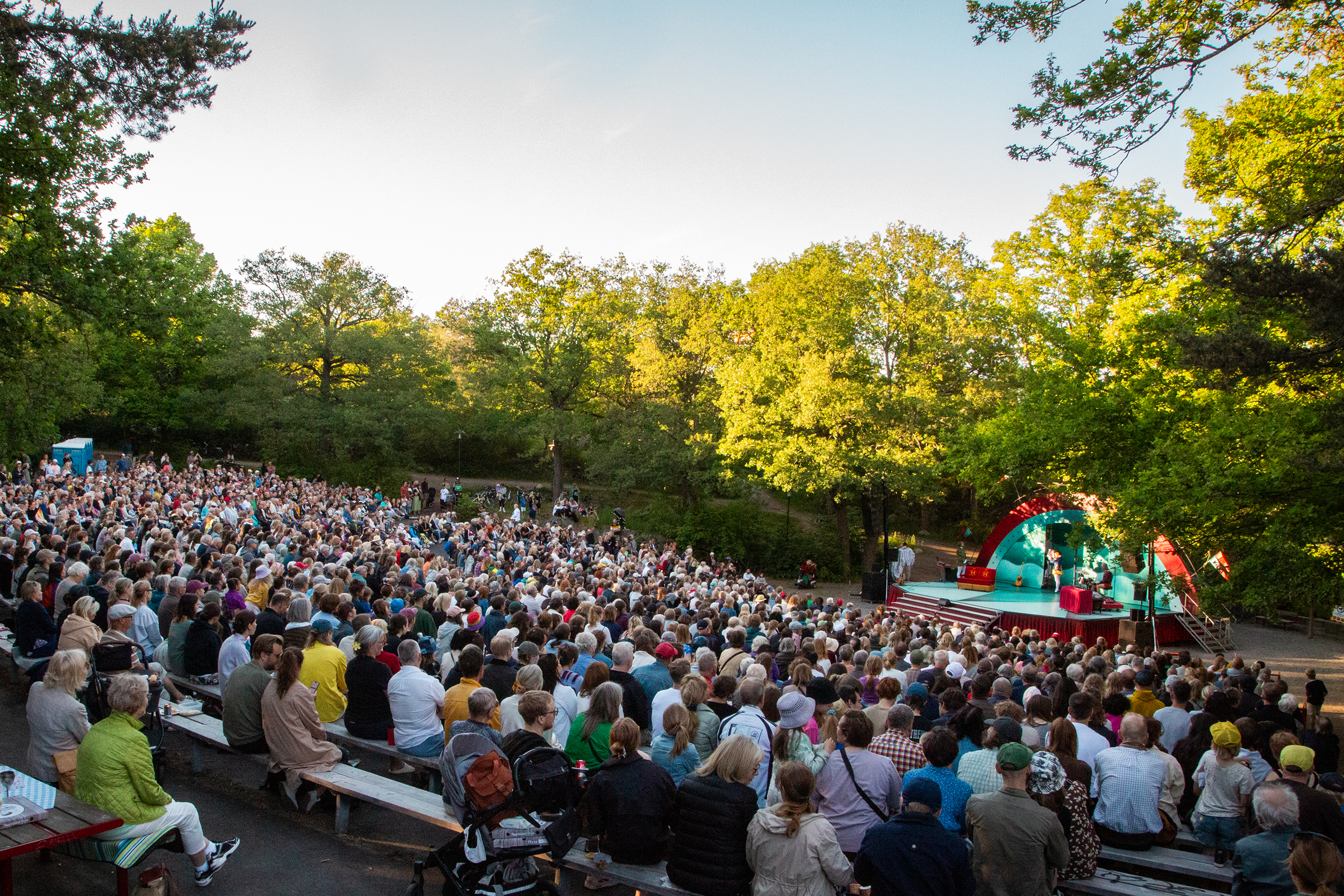 En stor publikgrupp som sitter framför en utomhusscen där det spelas teater en somrig kväll