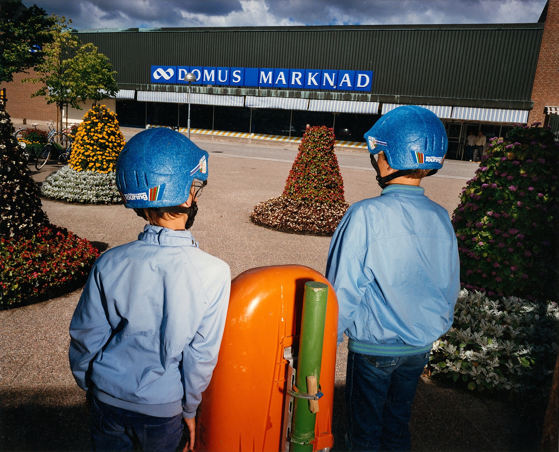 Två pojkar i blå hjälmar och jackor står vända mot varuhuset Domus Marknad, omgivna av blomstertorn.