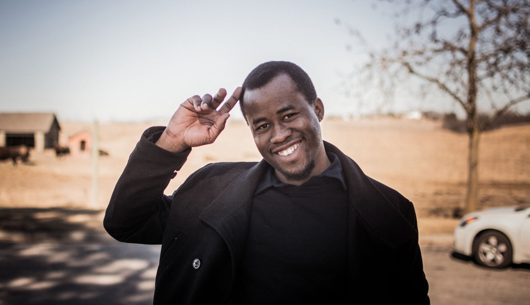 Ung man i mörk kappa och tröja, ler och gör honnör utomhus i lantlig miljö.