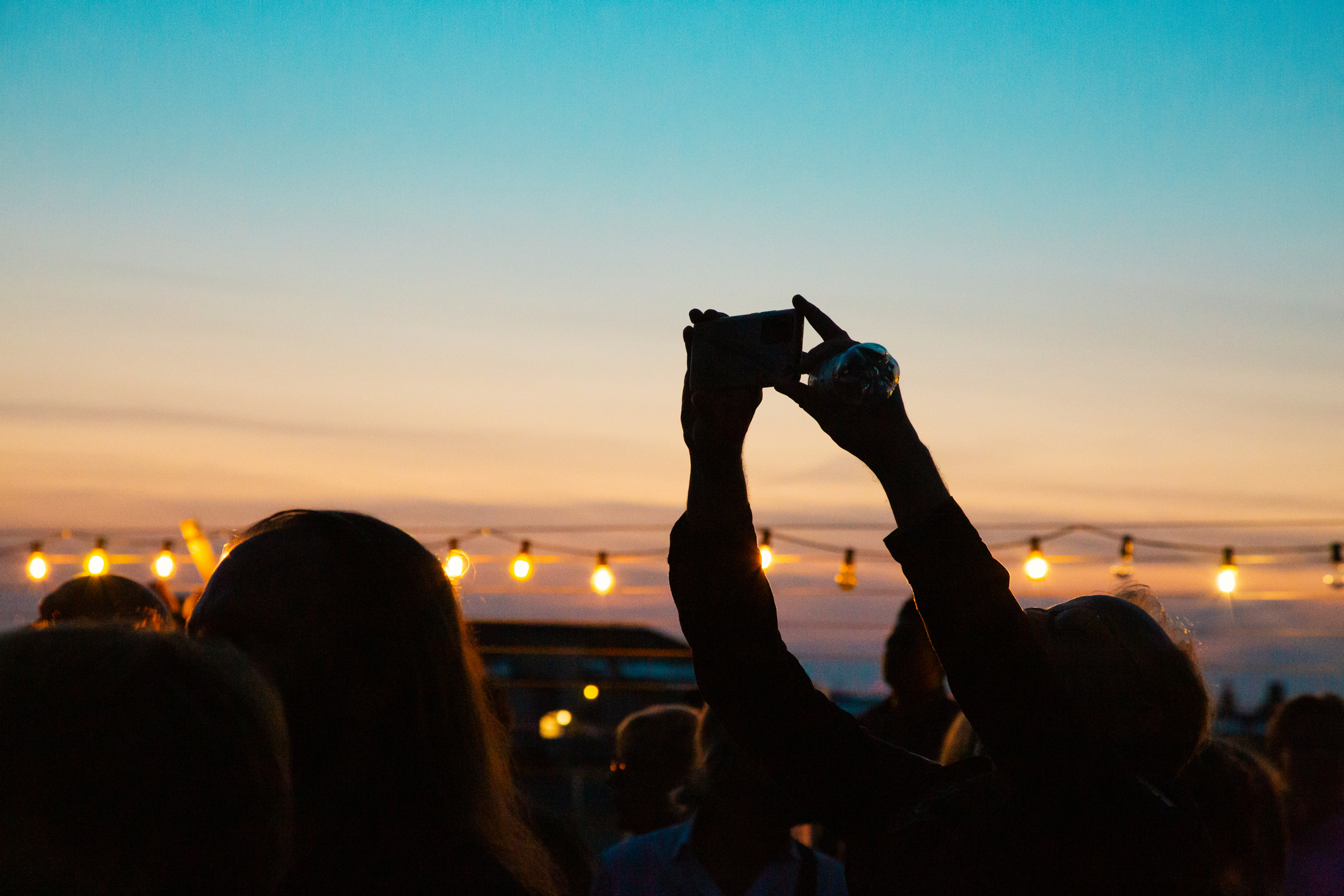 Silhuetter av personer i motljus vid solnedgång, en person håller upp en mobil mot himlen.