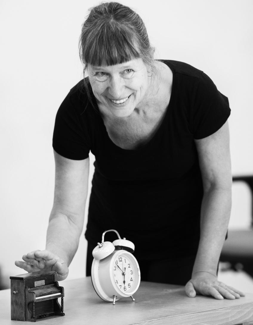 En kvinna står vid ett bord med en väckarklocka och ett miniatyrpiano på. 