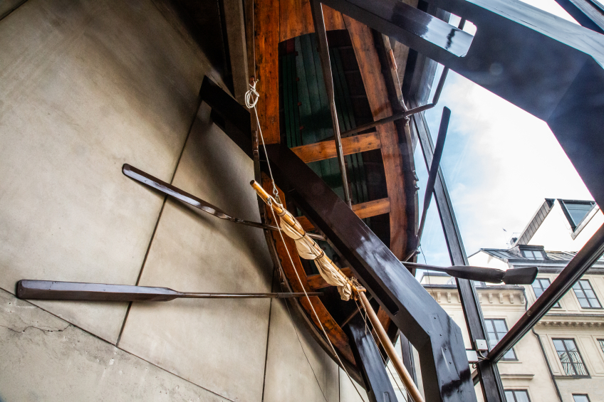 Ett konstverk som består av en båt och dess åror som hänger upp och ned i taket. En balk går rakt igenom båten.