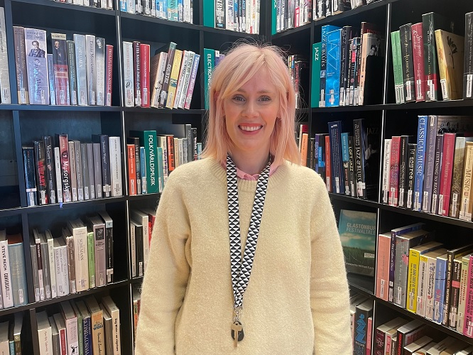 En bibliotekarie med rosa hår och ljus tröja står framför en hylla fylld med böcker på ett bibliotek och ler mot kameran.