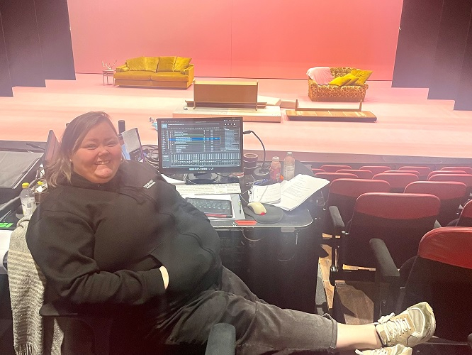 Kvinna som sitter avslappnat vid ett ljud- och ljusbord i en teater, med scenen i bakgrunden som är inredd med färgglada soffor och rosa bakgrund.
