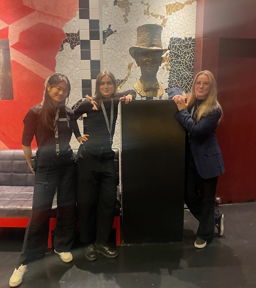 Tre personer poserar bredvid en staty, som föreställer en person med cylinderhatt. Rummet har färgglada väggar och mosaikdetaljer i bakgrunden.