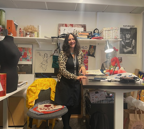 Sanna Tavares Nordenskjöld står vid sitt skrivbord i en ateljé, omgiven av tyg, modeillustrationer och en symaskin.