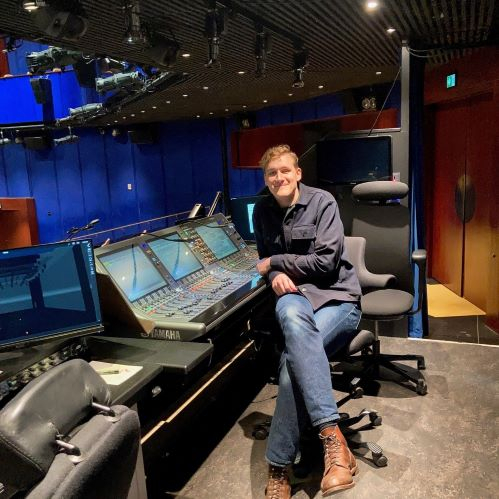 En man sitter avslappnat vid ett ljudbord i en studio, omgiven av teknisk utrustning och skärmar.