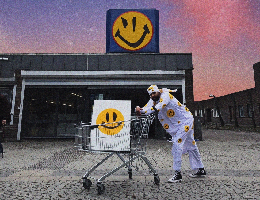 Unga man med kundvagn i vit smiley-dräkt framför buitk med drömsk himmel