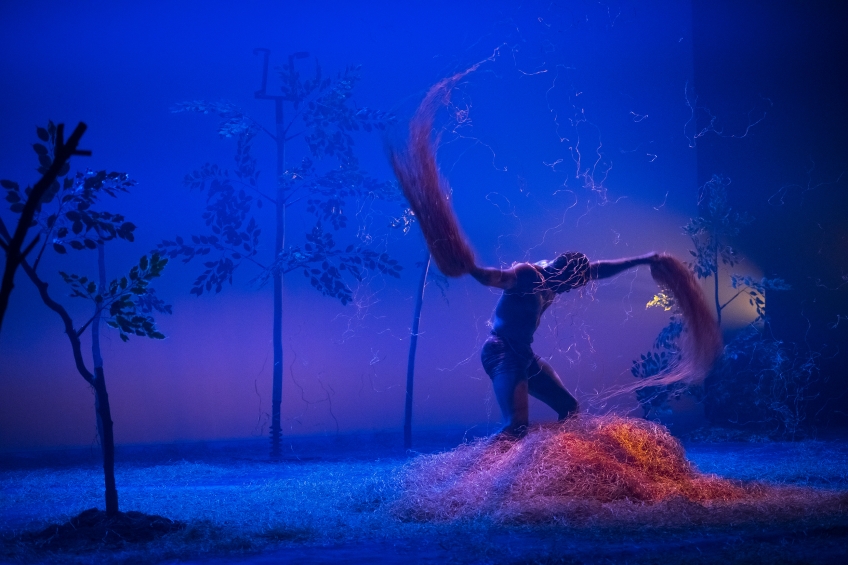 Person står på scen och kastar med armarna samtidigt som den håller i halm. Golvet är halmtäckt. Träd finns på scenen. Ljuset är blått.
