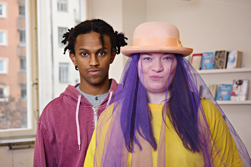 En tjej med hatt och ett lila genomskinligt tyg. Bakom henne står en kille