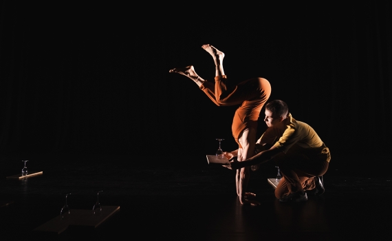 Kvinna står på händer med benen böjda framför en man som håller i en träplanka med ett vinglas på.