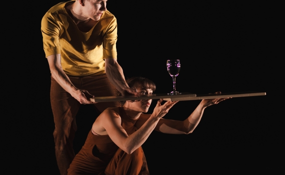 Svart bakgrund där kvinna står på knä och man står bakom, båda klädda i beige och bruna kläder. De håller i varsin träplanka som de använder för att hjälpa varandra balansera ett vinglas ovanpå.