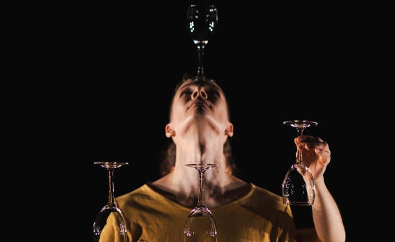 Man i beige t-shirt står mot svart bakgrund. Han håller i tre träplankor med 2 vinglas på. Ytterligare balanserar han ett vinglas på huvudet. En andra persons armar syns framför honom och håller i en träplanka med två vinglas på, samt håller ett tredje vinglas i luften.