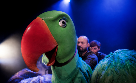 Två personer står bakom en stor docka som ser ut som en papegoja.
