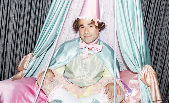 En man med struttat och kläder i turkost och rosa tittar snällt in i kameran. 