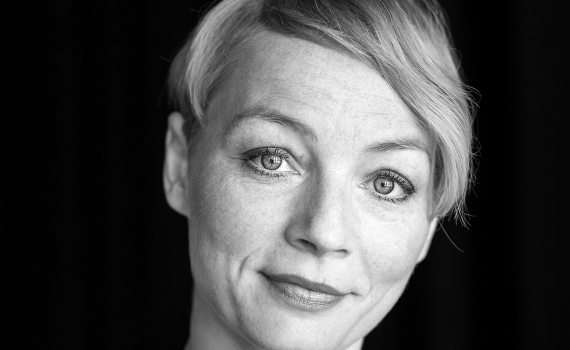 Porträtt av Sara Jangfeldt med kort ljust hår som ser rakt in i kameran med ett lugnt uttryck, svart bakgrund.