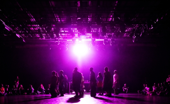 En grupp människor står i motljus på en scen med lila belysning, omgivna av sittande publik.