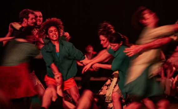 Dansare i färgglada kläder rör sig intensivt, en kvinna i grönt och orange dras av andra.