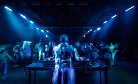 En DJ står vid ett bord med skivspelare, medan en suddig publik dansar under blå belysning.