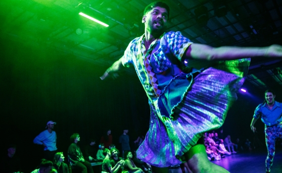 En man i mönstrad skjorta och glittrig kjol dansar, medan publiken sitter runt scenen.