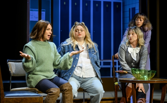 Fyra kvinnor sitter vid ett bord framför skåp, en av dem gestikulerar medan de andra lyssnar.