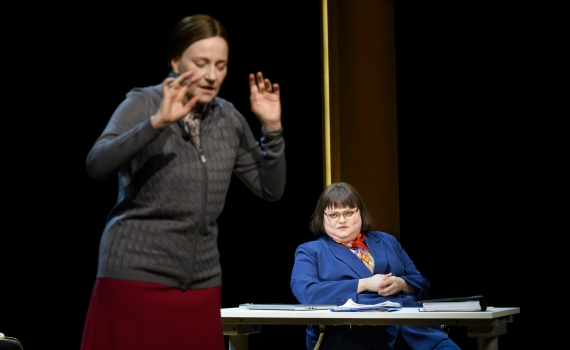 En kvinna i grå kofta gestikulerar, medan en annan i blå kavaj sitter vid ett skrivbord och tittar på henne.