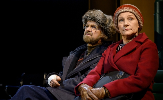 En äldre man i pälsmössa och en äldre kvinna i röd kappa sitter på en bänk och tittar åt sidan.