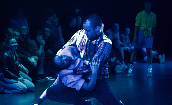 En man håller en kvinna i ett dramatiskt danslyft, omgivna av publik i blått ljus.