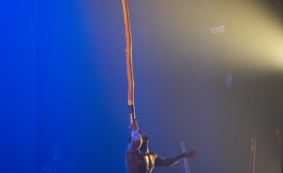 En man balaserar en lång pinne med en arm. Längst upp på pinnen hänger en annan man.