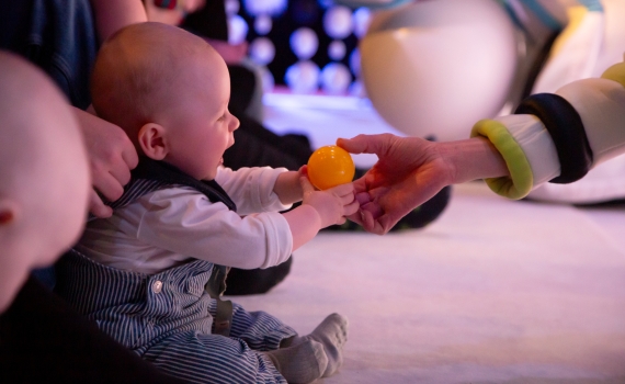 En baby i randiga hängselbyxor sitter på golvet och tar emot en gul boll från en person i vit och grön dräkt.