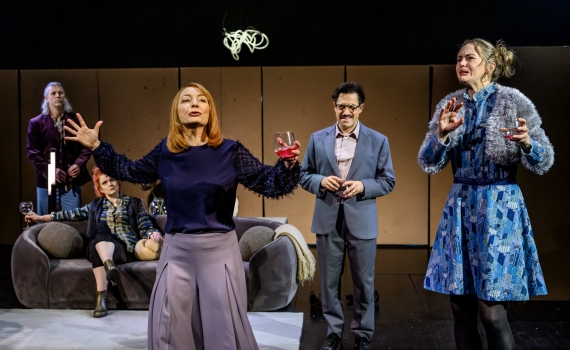 Fem vuxna skådespelare står på scen, två kvinnor i förgrunden med vinglas, en gestikulerar dramatiskt.