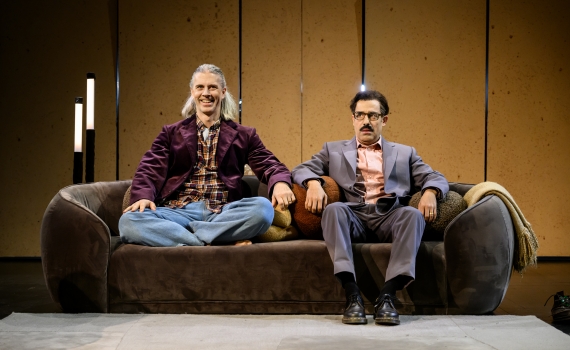 Två män sitter i soffa, den ene med långt hår och lila kavaj, den andre i grå kostym ser allvarlig ut.