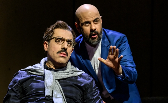 Två män sitter nära, en med glasögon och mustasch, den andre flintskallig i blå kavaj talar engagerat.