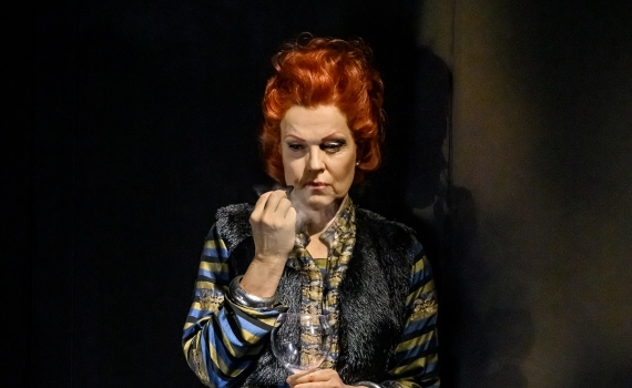 Kvinna med rött hår och randig dräkt står mot mörk vägg, håller ett glas och tittar ner.