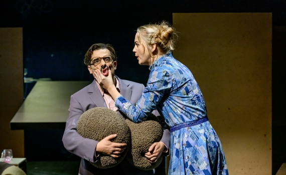 Kvinna i blå klänning håller hand mot man i kostyms ansikte, han håller en kudde mot kroppen.