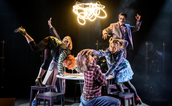 Sex vuxna agerar kaotiskt kring ett bord under ett stort lysande konstverk, mitt i en scen.