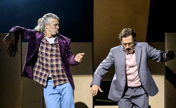 Två män hoppar ned från scenkonstruktion, den ene barfota i rutig skjorta, den andre i kostym.