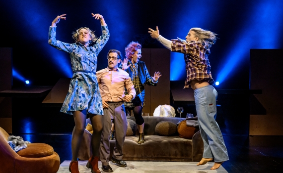 Fyra vuxna hoppar och gestikulerar på scen, kvinna i blå klänning och man i rutigt skjorta i rörelse.