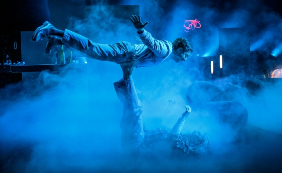 Man i ljus kostym balanserar horisontellt ovanpå en annan person, scenen är dimmig och blåbelyst.