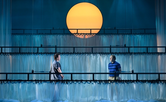 Två män står på varsin sida av en scen med balkongräcken och vit gardin, en stor sol lyser i bakgrunden.