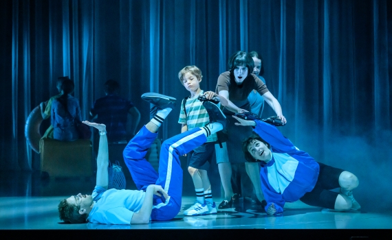 En pojke i randig tröja står bland vuxna och två personer i blå träningsoveraller som ligger på golvet i dramatisk scen.