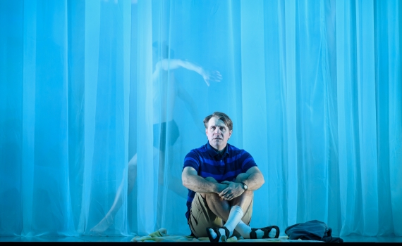 En man i blå pikétröja och shorts sitter på golvet framför blå genomskinliga draperier, en dansare syns bakom.