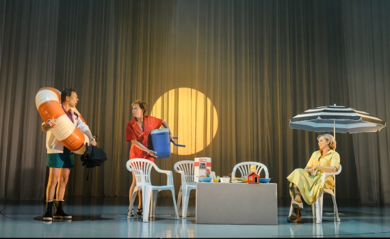 Två personer i badkläder och badleksaker står bredvid ett frukostbord, en kvinna i gul klänning sitter under parasoll.