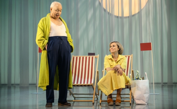 En äldre man i grön morgonrock står bredvid en kvinna i gul klänning som sitter i en solstol med en papperspåse bredvid.