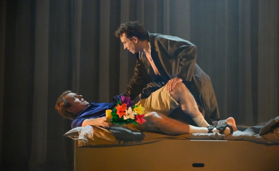En man i regnrock lutar sig över en liggande man med blommor i famnen på en sängliknande scenrekvisita.