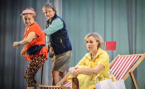 Två äldre kvinnor står vid en sittande yngre kvinna i gul klänning på en strandlik scen med solstolar.